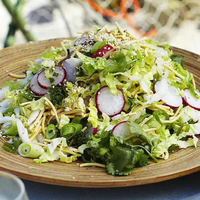 A wooden bowl filled with a fresh salad featuring sliced radishes, shredded lettuce, herbs, and sesame seeds.