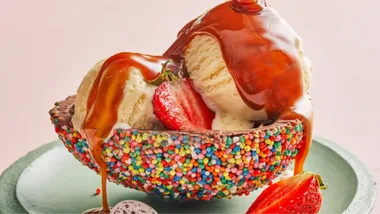 Ice cream scoops with caramel and strawberries in a sprinkle-covered bowl on a green plate.