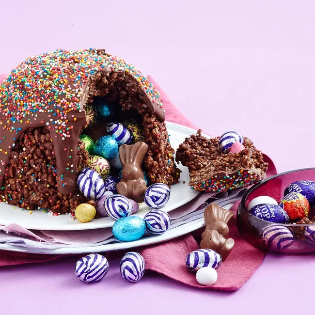 Chocolate egg dome with a sprinkle topping, filled with foil-wrapped candies and chocolate bunnies on a pink background.