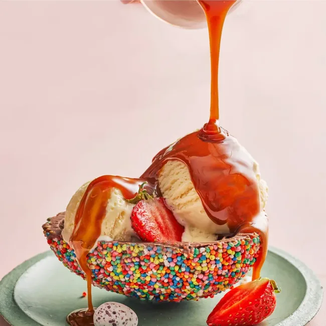 Ice cream scoops in a sprinkle-covered bowl, topped with caramel sauce and fresh strawberries.