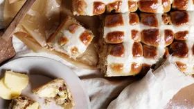 Freshly baked hot cross buns with a glaze, one partially eaten beside a plate with butter slices.