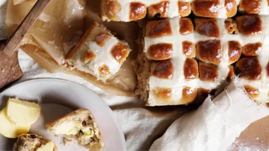 Freshly baked hot cross buns with a glaze, one partially eaten beside a plate with butter slices.