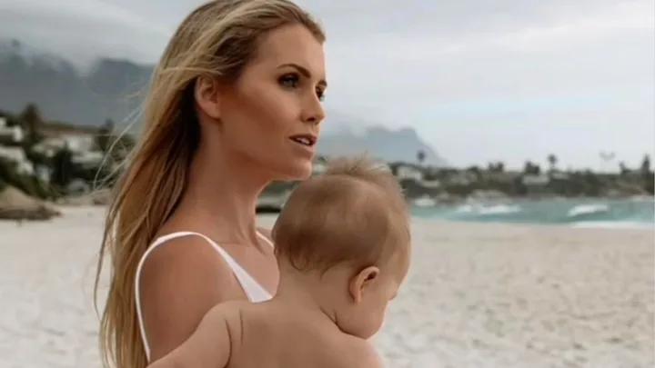 Woman holding baby on a beach, with mountains and ocean in the background.
