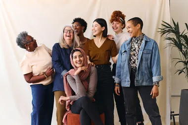 A diverse group of seven women laughing together against a neutral backdrop.
