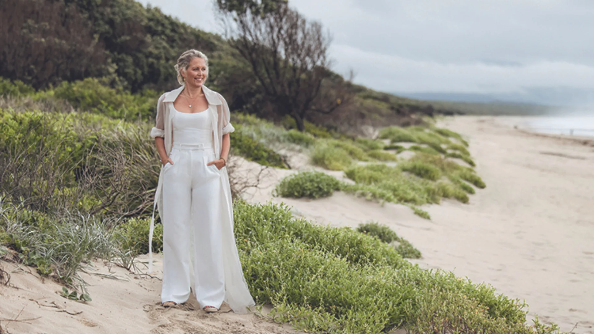 Woman in white outfit walking on a beach with greenery, cloudy sky.