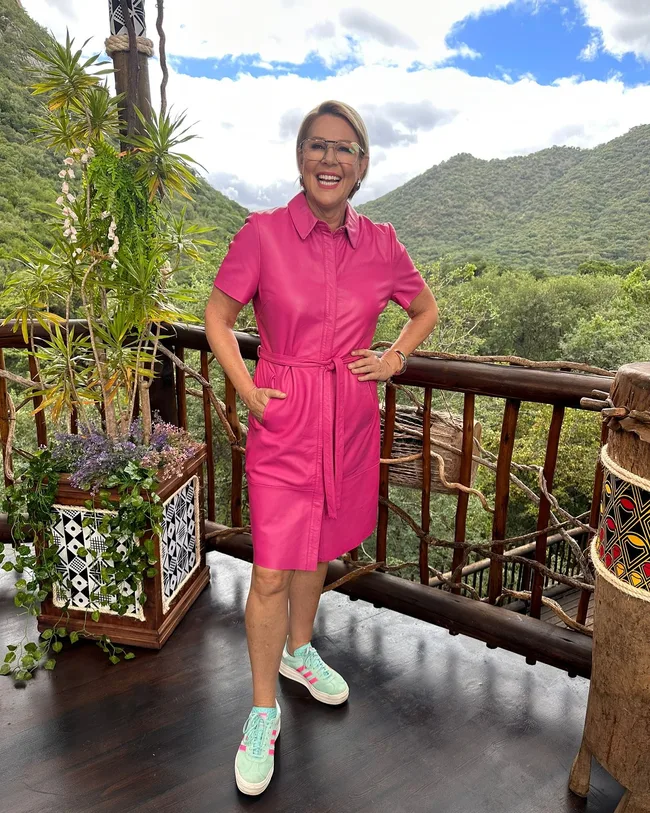 Woman in a pink dress and sneakers smiles on a wooden balcony, lush green hills and a cloudy sky in the background.