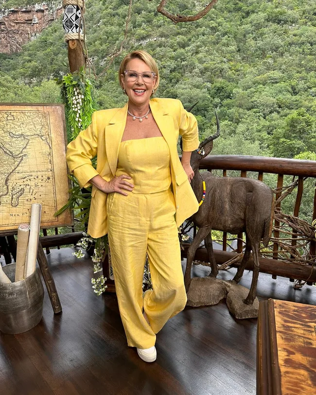 A woman in a yellow suit poses on a deck with lush greenery, a wooden statue, and an old map in the background.