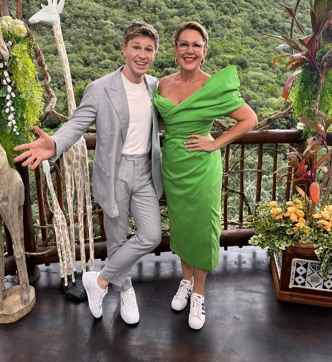 A man in a gray suit and a woman in a green dress pose on a balcony with a lush forest background.