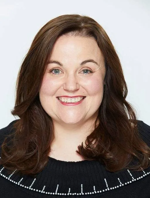 A woman with long brown hair smiling, wearing a black top with white patterns on the collar, against a plain background.