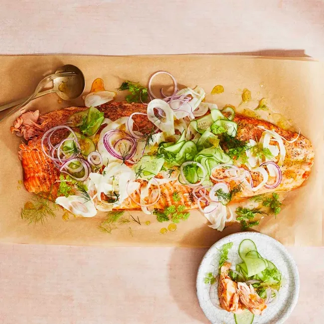 Grilled salmon topped with shaved fennel, cucumber, and red onions on parchment paper with a serving spoon nearby.