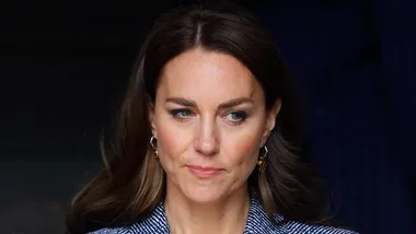 A woman with long brown hair and gold earrings, wearing a striped outfit, gazes downward.