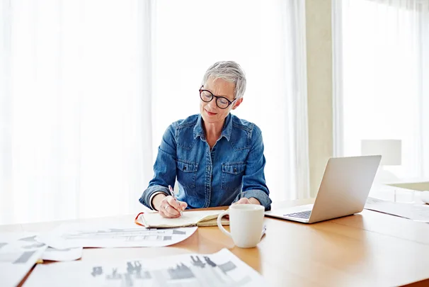 Elderly woman in glasses writing in a notebook at a desk with a laptop and papers, wearing a denim shirt.