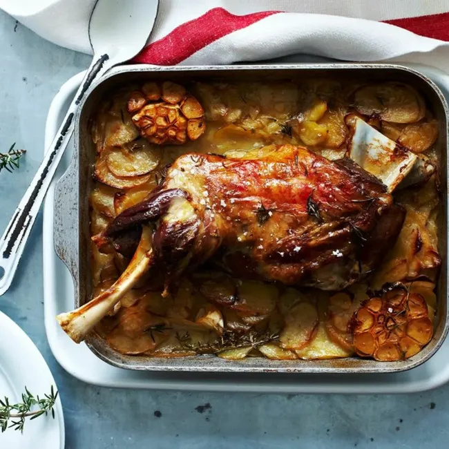 Roasted leg of lamb with garlic and rosemary in a baking dish, accompanied by sliced potatoes.