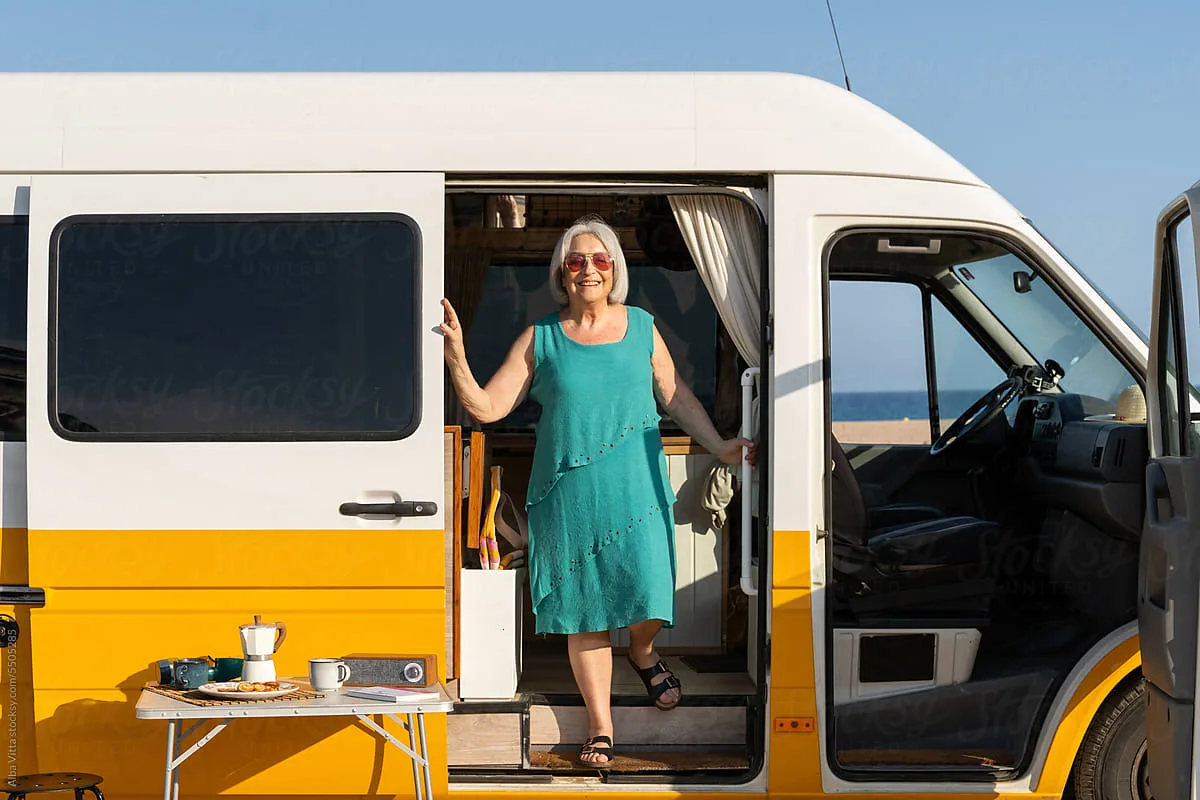 Smiling woman in teal dress stands at the open door of a yellow camper van by the ocean, with a table set for coffee.