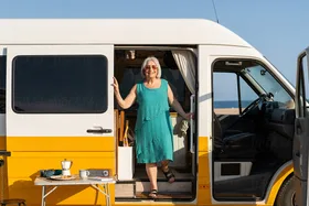 Smiling woman in teal dress stands at the open door of a yellow camper van by the ocean, with a table set for coffee.