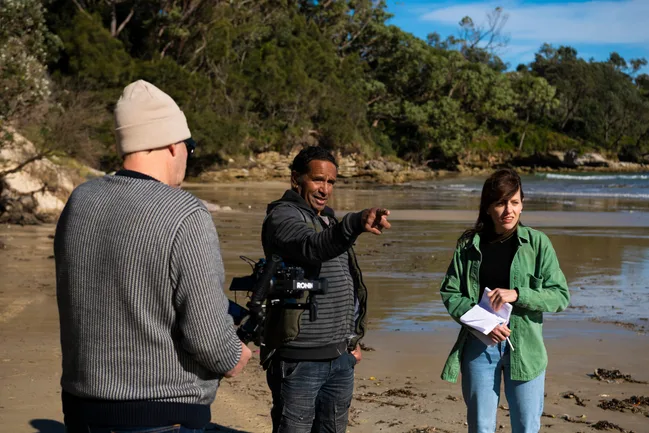 Film crew discusses scene on a sunny beach, featuring a camera and script in hand.