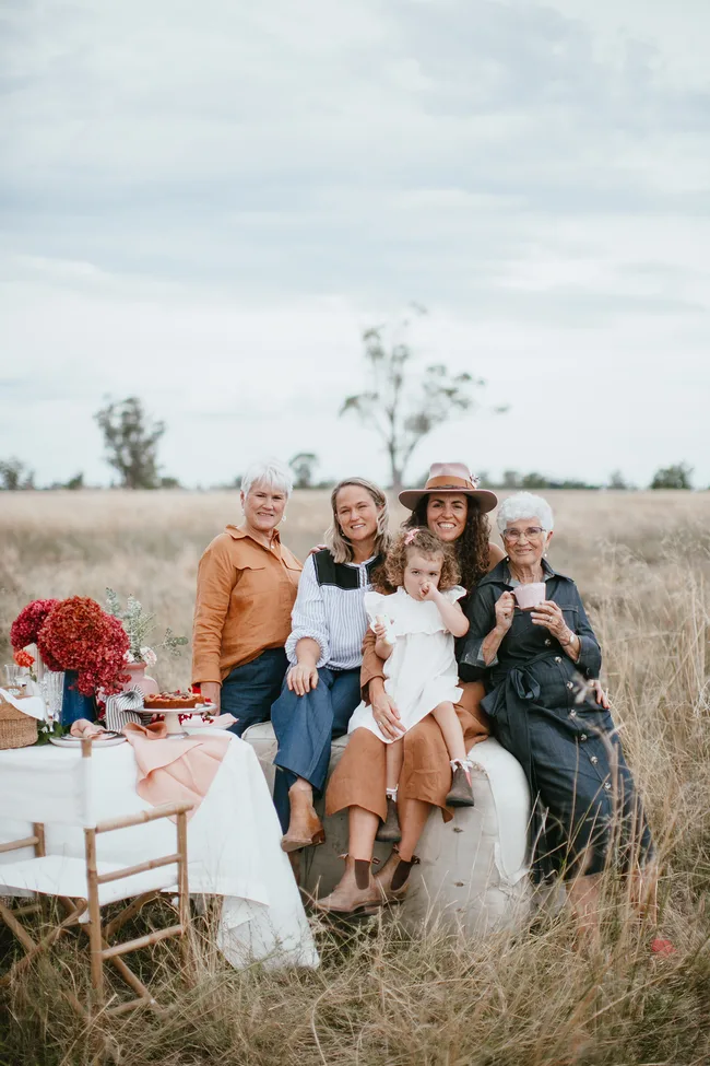 Family of five sits together on hay bale in a grassy field, smiling with a picnic setup beside them.