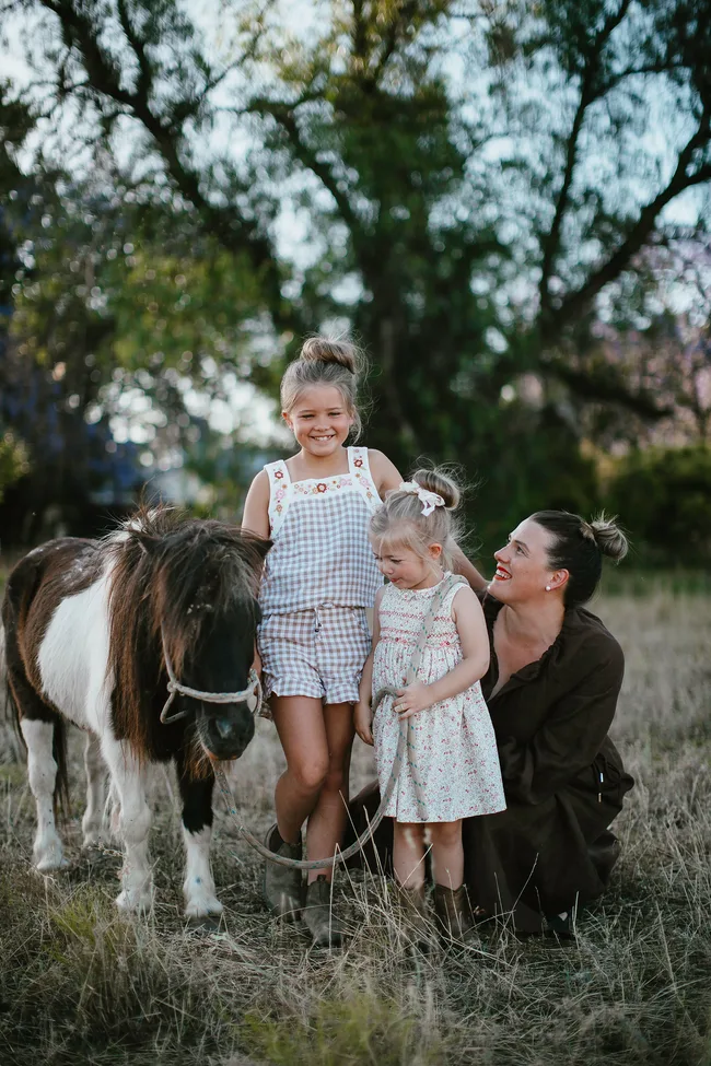 A woman kneels beside two young girls and a pony in a grassy field with trees in the background.