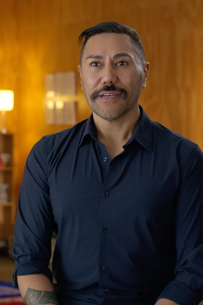 A person with a mustache and short hair, wearing a dark shirt, is speaking in a warmly lit room.