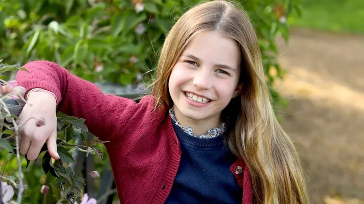 Young child with long hair smiles in a garden, wearing a red cardigan and dark top, surrounded by greenery.