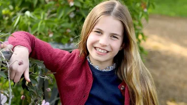 Young child with long hair smiles in a garden, wearing a red cardigan and dark top, surrounded by greenery.