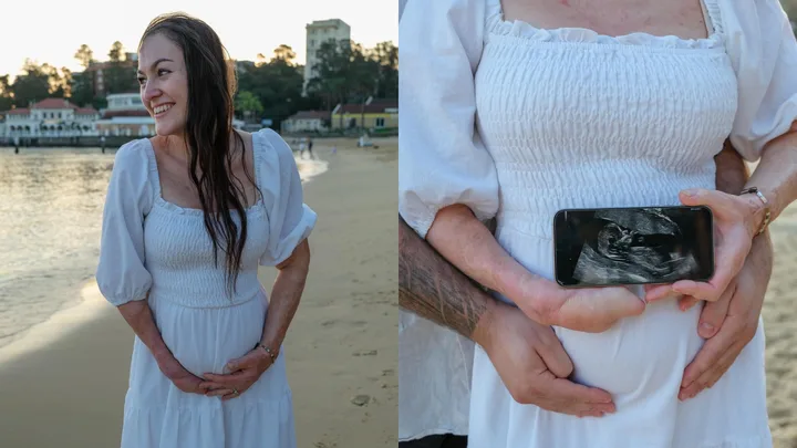 Pregnant woman in white dress smiles by the beach; phone shows ultrasound; partner hugs her from behind.