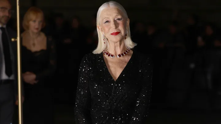 Helen Mirren in a sparkly black dress with a red necklace, posing on a red carpet event.