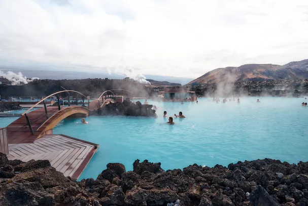 People enjoy the geothermal waters of Iceland's Blue Lagoon, with a wooden bridge and volcanic landscape in the background.