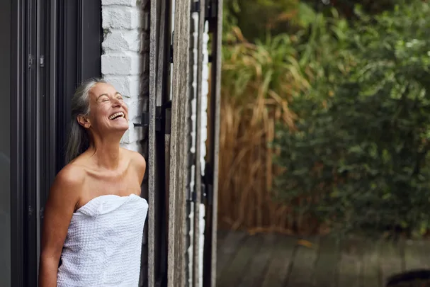 A woman wrapped in a towel laughs joyfully, standing by an open door with greenery outside.