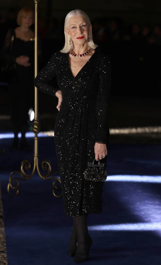 Helen Mirren in a sparkling black dress holding a small embellished bag on a blue carpet at a nighttime event.