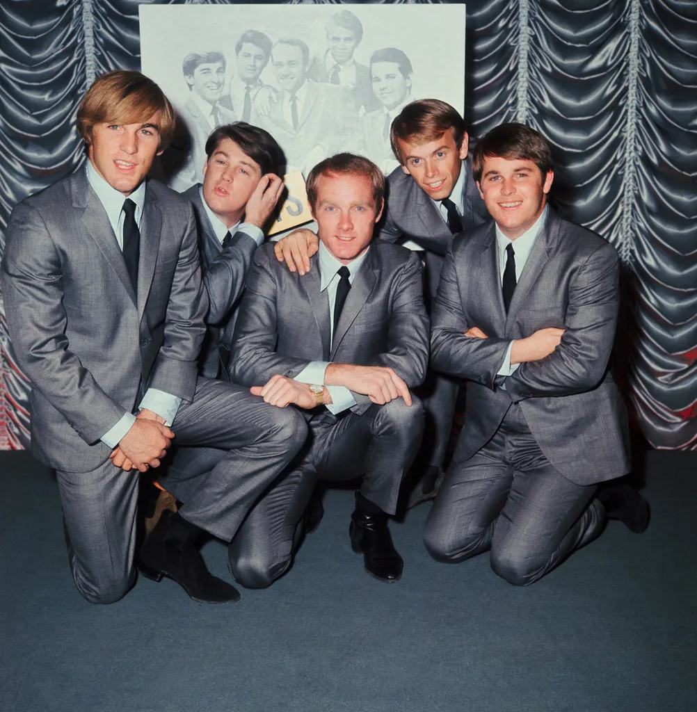 Five men in matching gray suits pose together, smiling, against a draped curtain background.