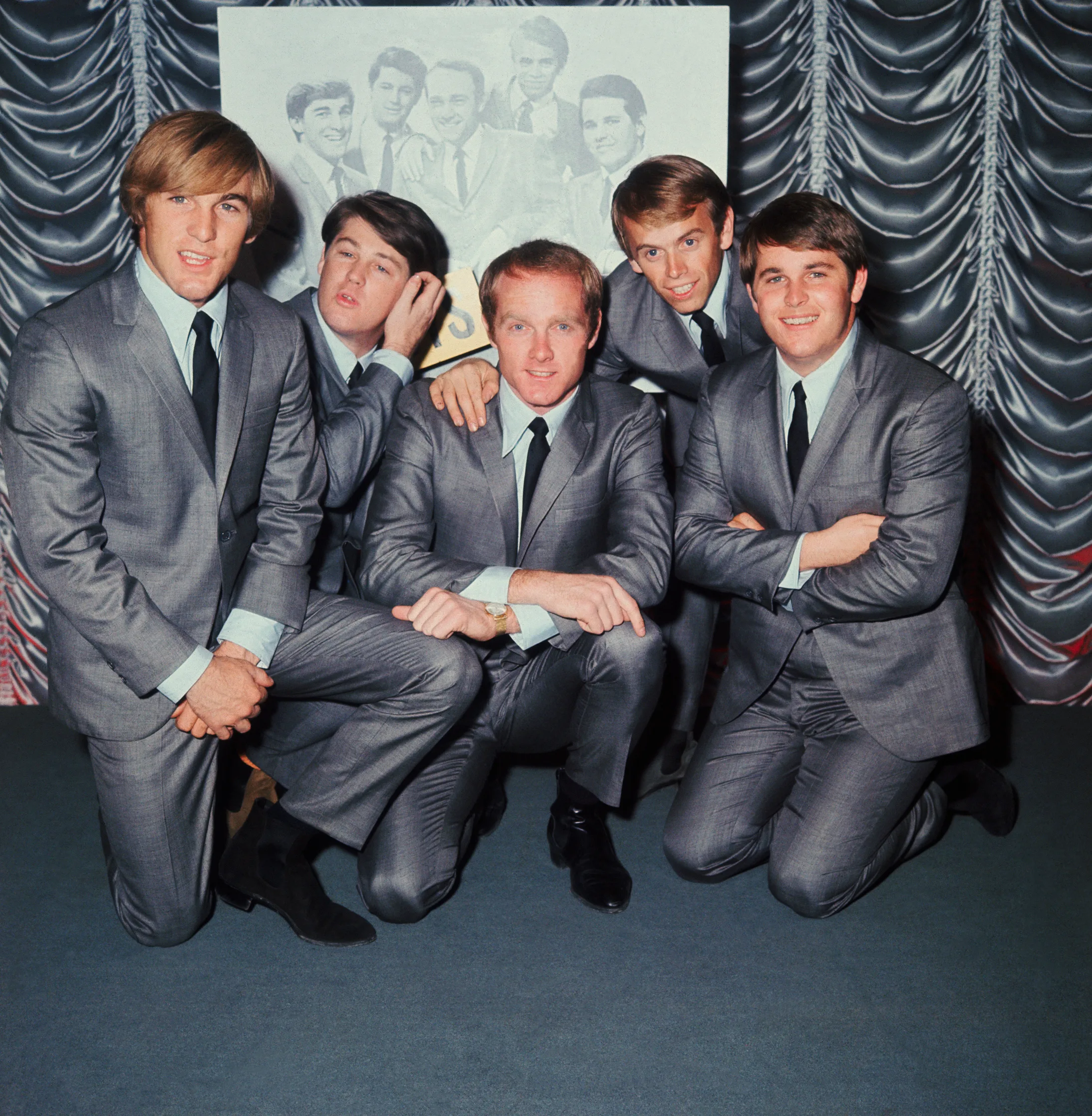 Five men in matching gray suits pose together, smiling, against a draped curtain background.