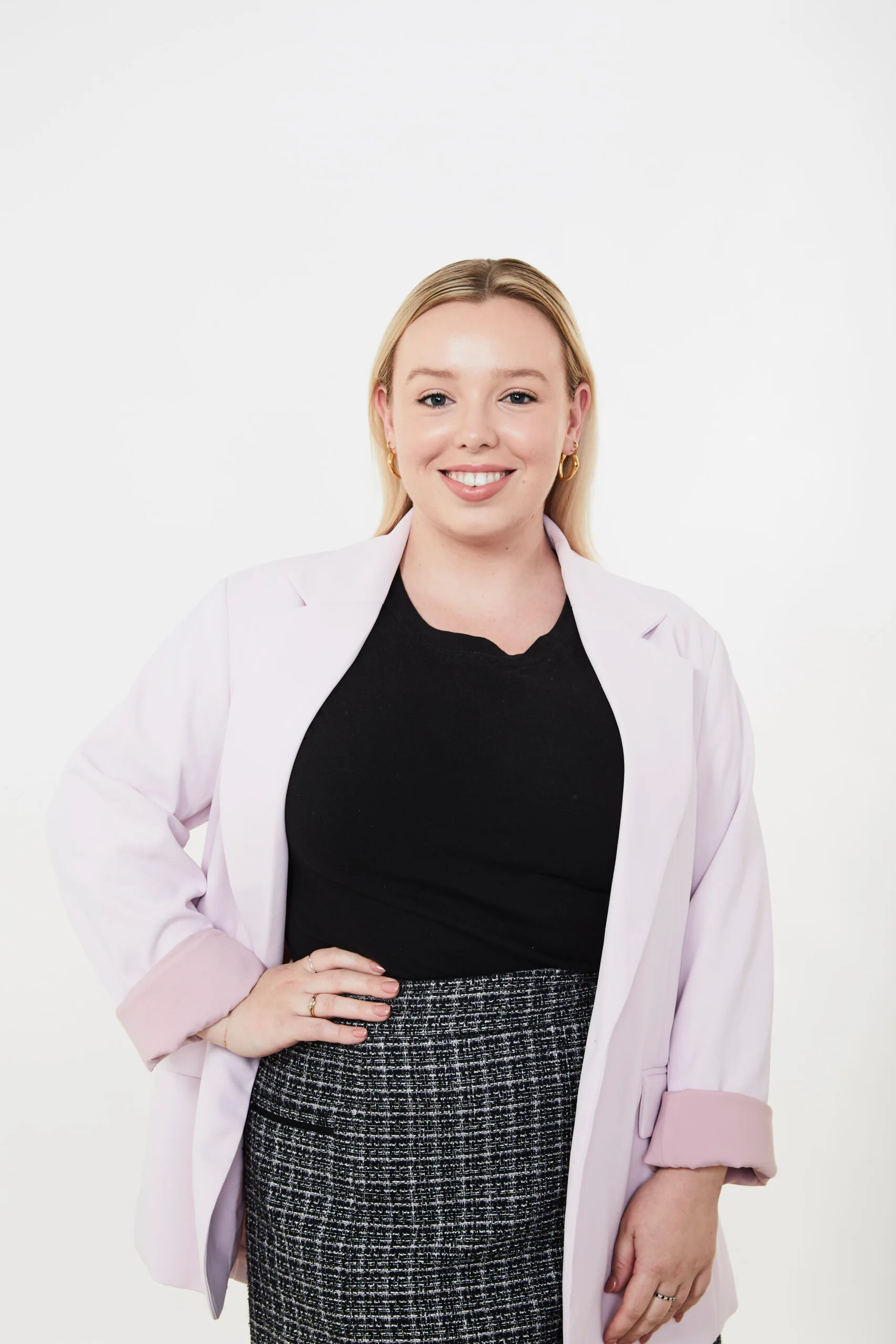 Smiling woman in a light purple blazer, black top, and checkered skirt poses against a plain white background.