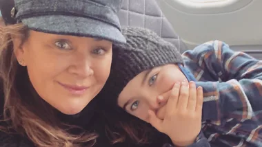 A woman in a cap and a child in a beanie and plaid shirt pose for a selfie inside a vehicle.