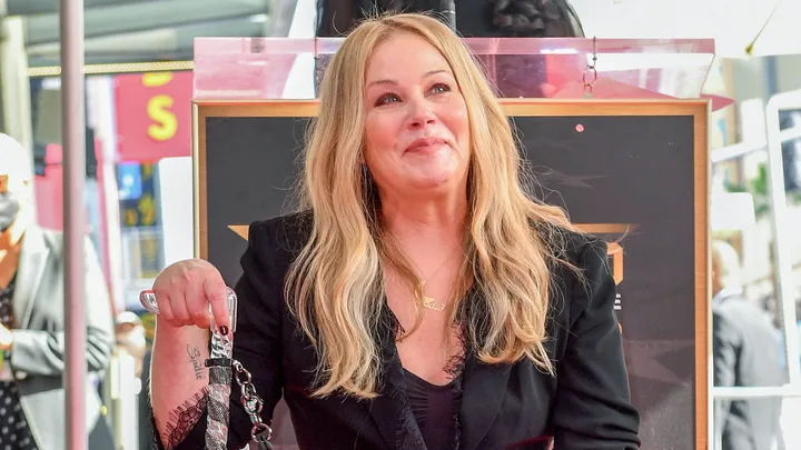 A woman with long blonde hair smiles while sitting at an event, wearing a black outfit and holding a walking aid.