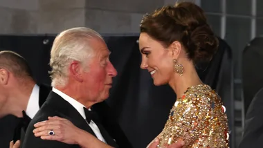 A man in a tuxedo and a woman in a gold dress smiling at each other at a formal event.