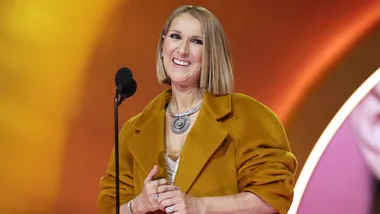 A woman in a mustard coat smiles while speaking into a microphone on stage with an orange background.