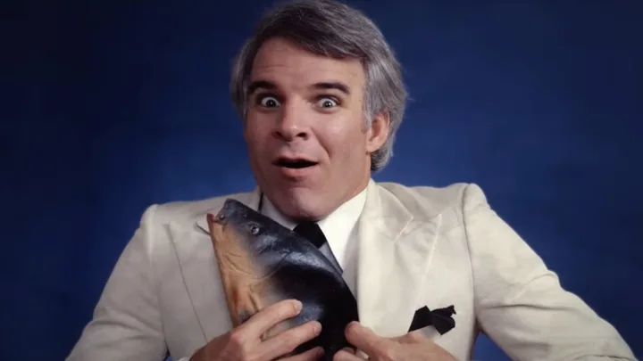Man in a white suit with surprised expression, holding a fish against a blue background.