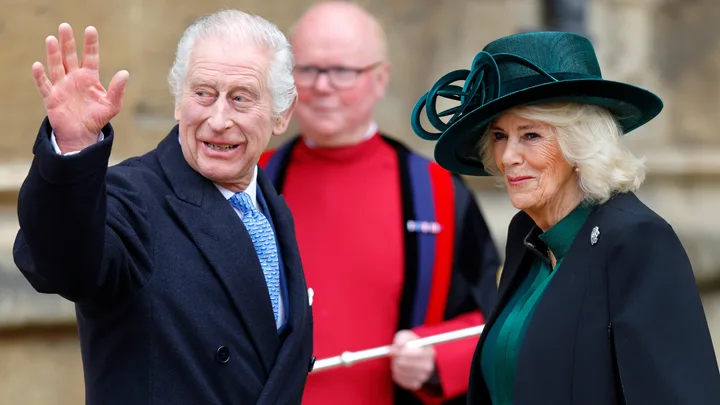 Elderly man waving, wearing a suit and blue tie, standing with a woman in a green hat and coat.