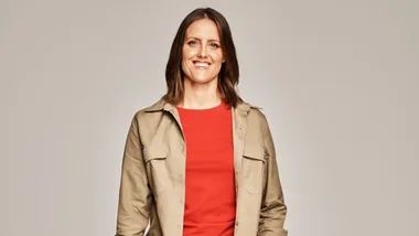 A woman with shoulder-length brown hair wearing a beige jacket and red top smiles at the camera against a neutral background.