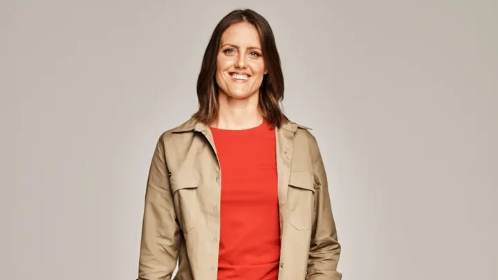 A woman with shoulder-length brown hair wearing a beige jacket and red top smiles at the camera against a neutral background.