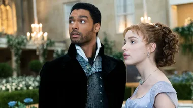 A man and woman in period costumes stand in an elegant ballroom, looking up with serious expressions.