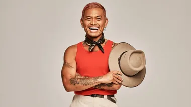 Man in red tank top with tattoos holding a hat, smiling broadly against a plain background.