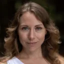 A person with wavy hair outdoors, wearing a white top, looking at the camera with a neutral expression.