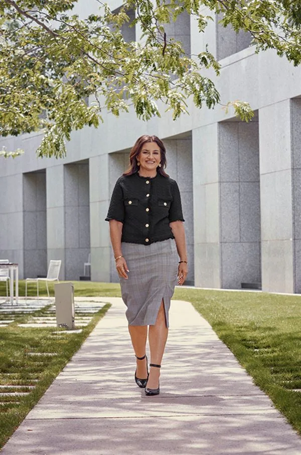 A woman in a black top and gray skirt walks on a path beside a modern building and trees under a clear sky.