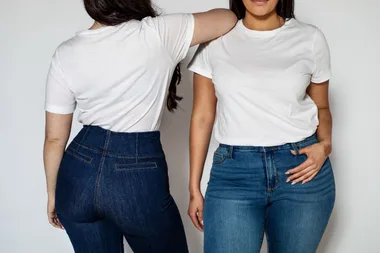 Two women in white t-shirts and blue jeans, one facing forward and the other turned, standing against a neutral background.