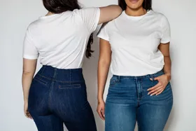 Two women in white t-shirts and blue jeans, one facing forward and the other turned, standing against a neutral background.