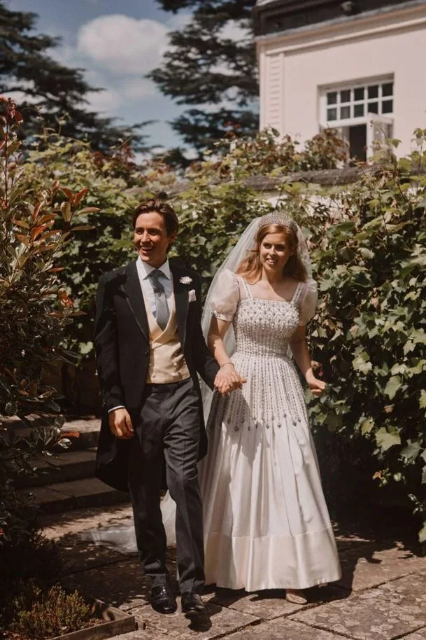 Bride in a white gown and groom in a morning coat walking outside, surrounded by greenery on a sunny day.