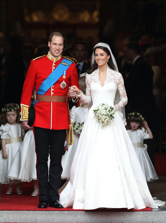 Bride in lace gown and groom in red uniform, smiling as they leave church with flower girls.