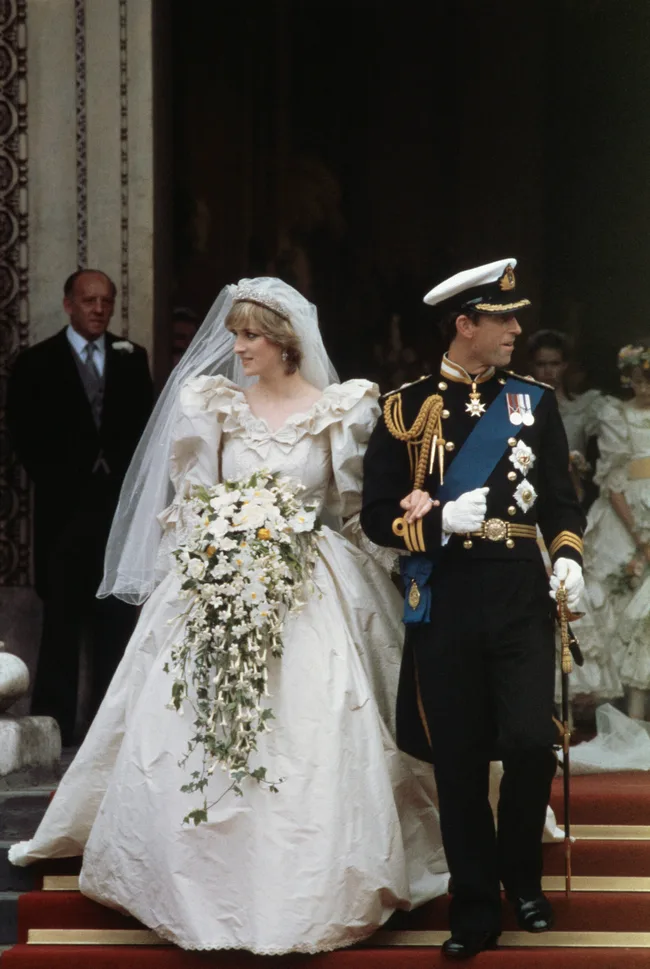A bride in an ornate gown with a long veil and floral bouquet walks hand-in-hand with a uniformed groom down stairs.